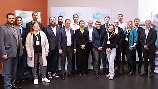 18 Personen stehen aufgereiht mit Blick in die Kamera