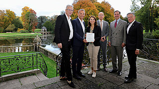 Sechs Personen stehen vor einem Schlossteich auf einer Terrasse. Zwei Personen halten gemeinsam eine Urkunde