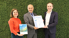 Drei Personen stehen vor einer grünen Mooswand und halten die EMAS-Urkunde und die ZUG-Umwelterklärung in die Kamera.