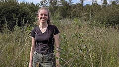 Eine Frau steht auf einer Grünfläche und lächelt, im Hintergrund Bäume 