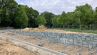 Steinmauer in einem Baustellenbereich mit Sandhaufen und Bäumen im Hintergrund