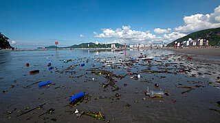 Strand mit verstreutem Plastikmüll und Treibgut im Vordergrund, dahinter Stadt und bewaldete Hügel unter blauem Himmel mit Wolken