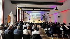 Großer Saal mit Publikum, das auf eine Präsentation an einer Leinwand in einem Raum mit hohen Fenstern und hängenden Lampen blickt