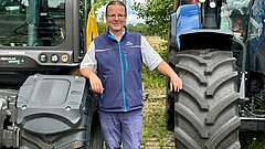 Mann mit Brille und blauem Weste lehnt an großen Reifen von zwei landwirtschaftlichen Fahrzeugen im Freien.