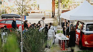 Mehrere Personen stehen vor einem Pavillon, unter dem Pavillon steht ein Tisch, und die Personen sehen sich an, was auf dem Tisch liegt. 