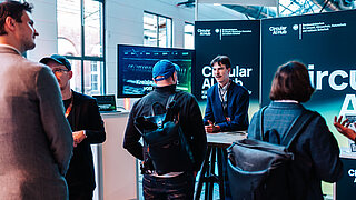 Mehrere Personen unterhalten sich vor einem Messestand mit Bildschirmen und Bannern des Circular AI Hub in einem hellen Raum mit großen Fenstern.
