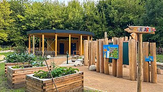 Blick auf Hochbeete, einen Pavillion und mehrere in den Boden eingegrabene Stämme mit Bildtafeln und Schildern