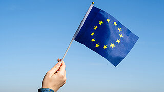 Eine Hand hält eine kleine Fahne mit EU-Flagge vor blauem Himmel