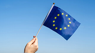 Eine Hand hält eine kleine Fahne mit EU-Flagge vor blauem Himmel