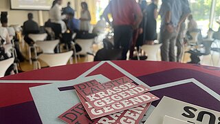 Flyer im Vordergrund; Teilnehmende beim Pitch im Hintergrund