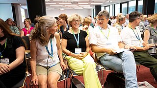 Mehrere Personen sitzen in Stuhlreihen und sprechen miteinander. 