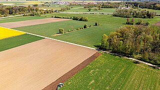 Luftaufnahme einer landwirtschaftlich geprägten Landschaft mit Feldern, Wiesen und Baumgruppen. Im Vordergrund bearbeitet ein Traktor den Rand eines Ackers; im Hintergrund sind weitere Felder und eine Ortschaft zu sehen.