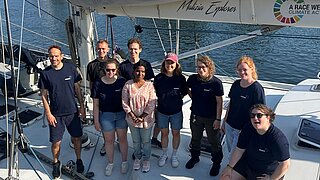 Neun Personen stehen auf dem Deck eines Segelbootes
