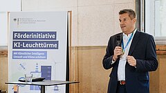 Ein Mann (Thorsten Scholz) steht neben einem Rollup und spricht in ein Mikrofon