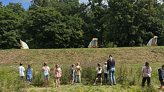 Kinder und eine erwachsene Person stehen auf einer Wiese vor einem Erdwall mit drei kleinen Segelbootmodellen