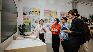 Einige Personen stehen vor einem Bildschirm und schauen sich etwas an, eine weitere Person erklärt etwas dazu. 