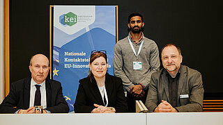 Drei Personen sitzen an einem Tisch. Ein vierte steht dahinter neben einem Roll-up mit dem Logo des KEI.