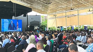 Ein großes Pubikum sitzt vor einem Bildschirm in einer Messehalle. Auf dem Bildschirm ist ein Mann zu sehen
