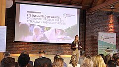 Eine Frau (Anna Hecker) steht auf einer Bühne. Hinter ihr eine Projektionsleinwand, vor ihr das Publikum