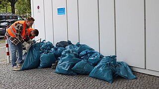 Diverse Müllsäcke liegen an einem Gebäude. Daneben stehen 2 Personen