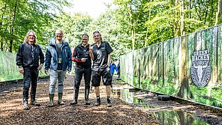 Vier Personen (drei in Gummistiefeln) stehen neben einem Zaun auf dem "Green Wacken" steht. 