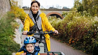 Eine Frau sitzt auf einem Lastenfahrrad und fährt durch eine grüne, städtische Region. Vor ihr sitzt ein Kind mit Helm. 