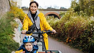 Eine Frau sitzt auf einem Lastenfahrrad und fährt durch eine grüne, städtische Region. Vor ihr sitzt ein Kind mit Helm. 