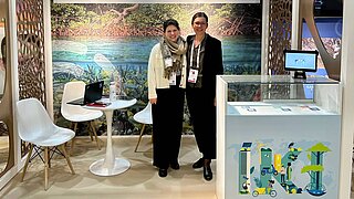 Zwei Personen stehen vor einem Messestand mit Thresen, Tischen und Stühlen. Auf dem Thresen und im Hintergrund sieht man das IKI Logo