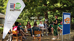 Mehrere Personen sitzen auf Stühlen im Freien, eine Person steht, neben ihnen eine Pinnwand und eine Beachflag