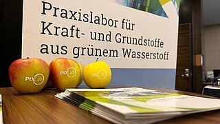 Broschüren und Äpfel auf einem Tisch, im Hintergrund ein Poster