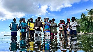 Eine Gruppe Menschen steht im Meer, ihre Spiegelbilder werden im Wasser reflektiert, im Hintergrund Wolken und ein Palmenstrand. 