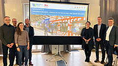 Sieben Personen stehen rechts und links neben einem großen Bildschirm. 