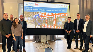 Sieben Personen stehen rechts und links neben einem großen Bildschirm. 