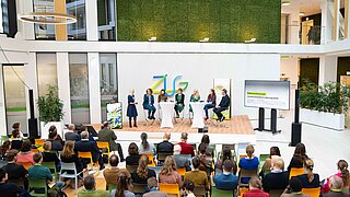 Auf einer Bühne sitzen Personen und diskutieren, im Vordergrund befindet sich das Publikum