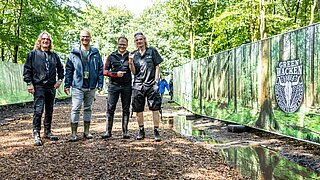 Vier Personen (drei in Gummistiefeln) stehen neben einem Zaun auf dem "Green Wacken" steht. 