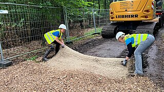 Zwei Personen mit Schutzhelmen legen Paludi-Matten auf den Boden und bedecken sie mit Holzschnitzeln. Im Hintergrund steht ein Baufahrzeug. 