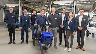 Eine Gruppe von 8 Personen in einer Lagerhalle, die mittlere Person sitzt auf einem Lastenrad. 