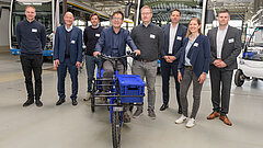 Eine Gruppe von 8 Personen in einer Lagerhalle, die mittlere Person sitzt auf einem Lastenrad. 