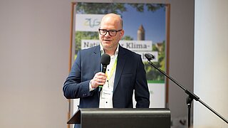 Ein Mann steht an einem Rednerpult und hat ein Mikro in der Hand, hinter ihm sieht man ein Poster 