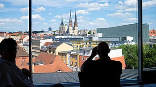 Blick aus einem Fenster auf die Stadt Halle. Am Fenster stehen zwei Personen und sehen hinaus. 