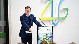 Ein Mann (BM Carsten Schneider) steht an einem Redepult, im Hintergrund das Logo der ZUG