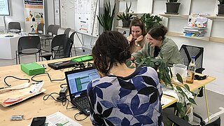 Drei gut gelaunte Frauen sitzen an einem Tisch vor Computern und Technik