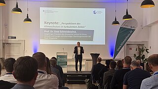 Vortragender steht auf Bühne vor Publikum in hellem Saal mit Leinwand und Banner zum Thema Klimaschutz