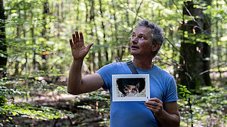 Sprechender Mann steht im Wald und zeigt ein Foto.