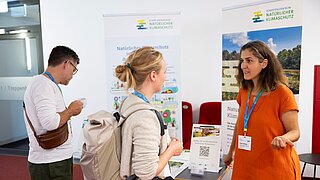 Drei Personen befinden sich an einem Messestand, reden miteinander und schauen sich Materialien auf einem Tisch an