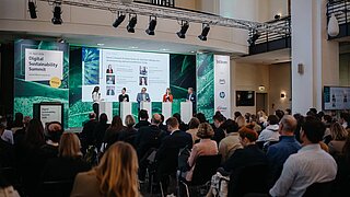 im Hintergrund eine Bühne mit Sprechenden, im Vordergrund das Publikum 