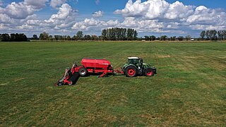Luftaufnahme eines grünen Traktors mit roter Sämaschine auf einer weitläufigen Grünlandfläche. Im Hintergrund sind Baumreihen unter einem wolkigen Himmel zu sehen.