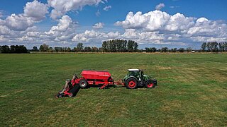 Luftaufnahme eines grünen Traktors mit roter Sämaschine auf einer weitläufigen Grünlandfläche. Im Hintergrund sind Baumreihen unter einem wolkigen Himmel zu sehen.