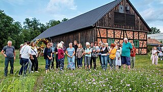Mehrere Personen stehen auf einer blühenden Wiese, im Hintergrund ein Fachwerkgebäude