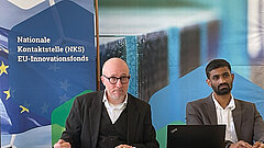 Zwei Personen - Frank Paul Weber und Vinay Reddy Kambam - sitzen vor einem Banner mit der Aufschrift "Nationale Kontaktstelle (NKS) EU-Innovationsfonds, ein Laptop
